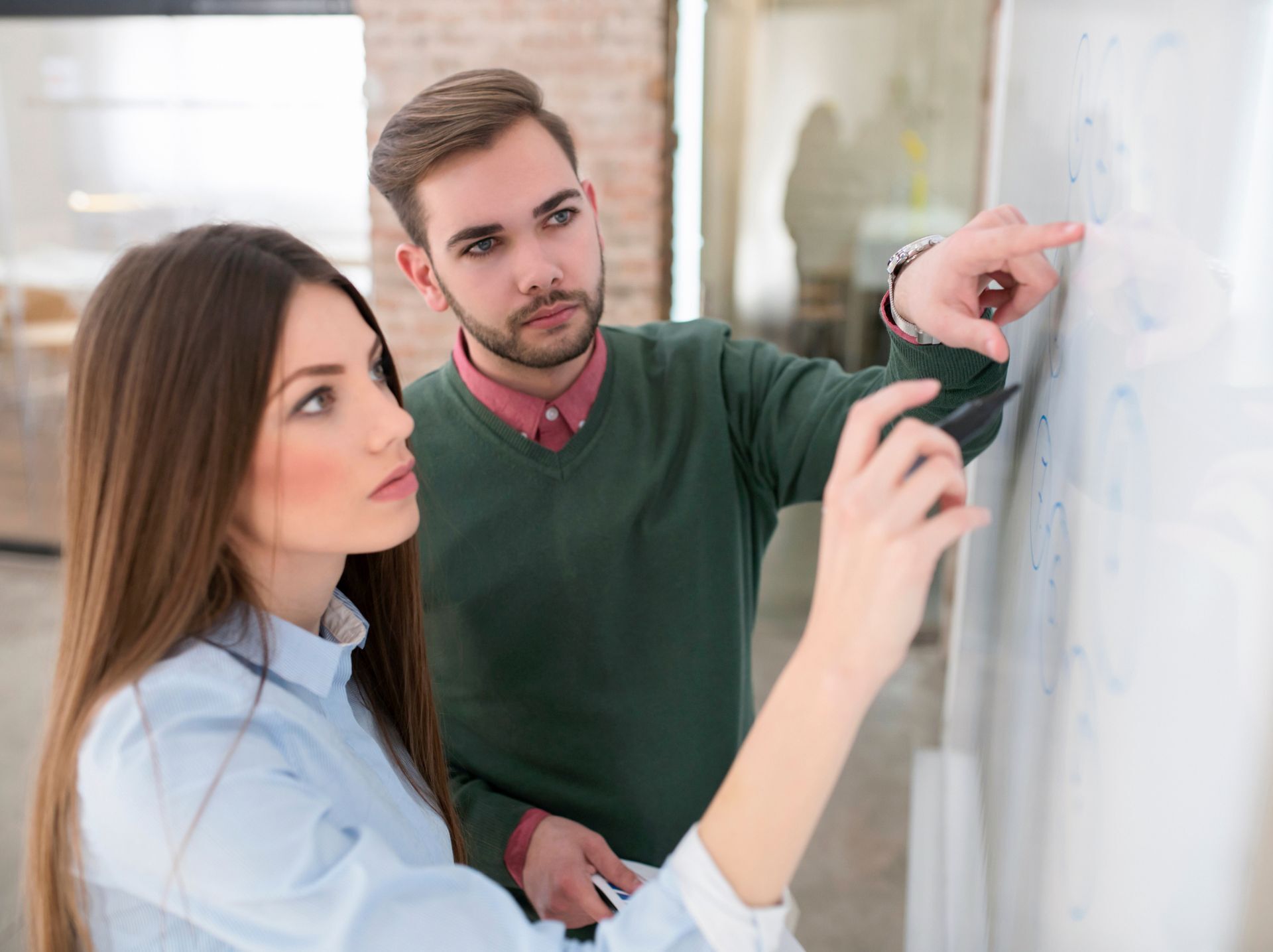 A man and a woman in an office collaborate at a whiteboard. The woman writes or points, while the man observes and points, discussing marketing forecasting and trends.