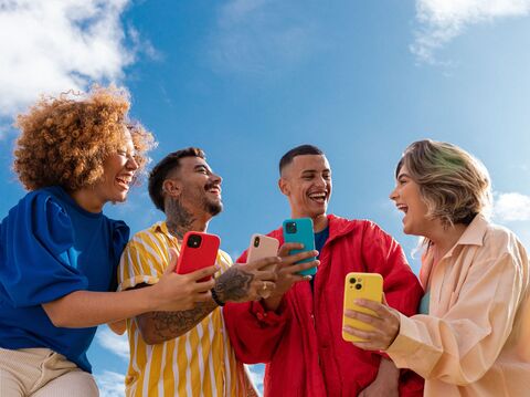 Four joyful young adults, diverse in ethnicity, stand together outdoors under a bright blue sky, each holding a smartphone. They are engaging with each other and their devices, sharing a light-hearted moment