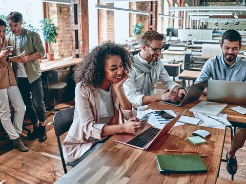 A diverse group of young professionals collaboratively working in a stylish, spacious office. One woman with curly hair smiles while using her smartphone, sitting beside two men focused on laptops. Another male and female pair stand discussing over a smartphone