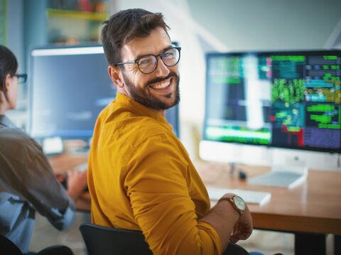 A person wearing glasses and a yellow shirt surrounded by screens and lines of code