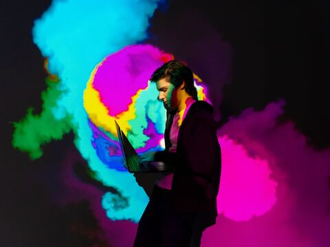 A young man with dark hair, working on a laptop against a vivid backdrop of swirling, multicolored neon clouds