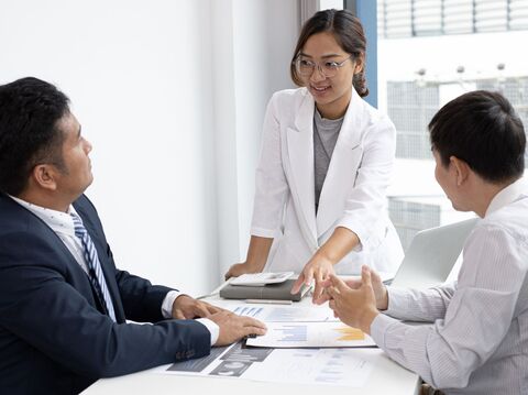 A woman in a light-colored blazer presenting charts to two male colleagues in a bright office setting