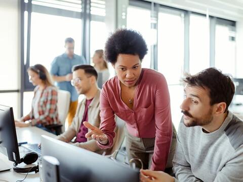 six people in the frame two in the background chatting two on their workstation and two discussing something on the screen