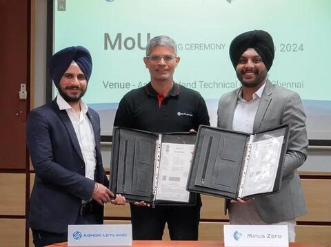 Three executives from Ashok Leyland and Minus Zero holding an agreement folder at a MoU signing ceremony in Chennai. They are standing in front of a projector screen displaying 'MoU Signing Ceremony 2024