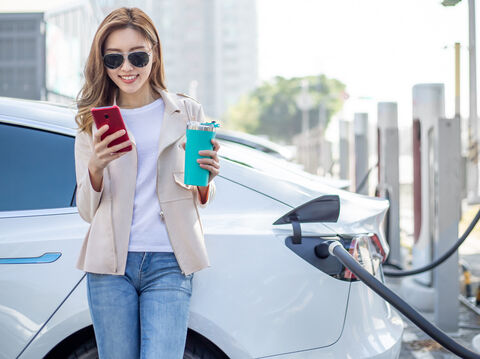 A woman is standing in front of an electric car, holding a sipper in one hand and a smartphone in the other. The electric car is getting charged.