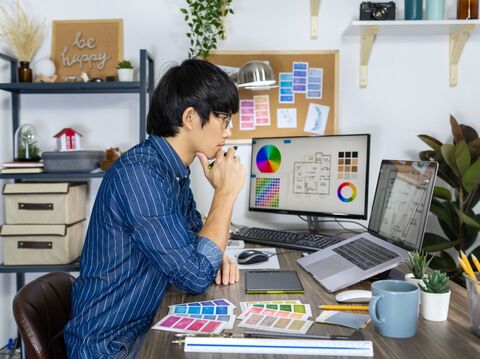 A young man deep in thought, sitting at a cluttered desk surrounded by various design tools and color palettes, with a computer screen displaying graphic charts and design layouts