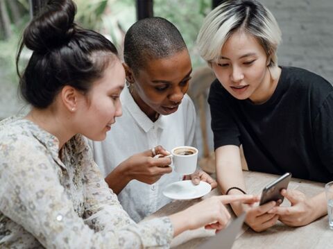 Three diverse women, looking at a smartphone and discussing something together at a cafe table