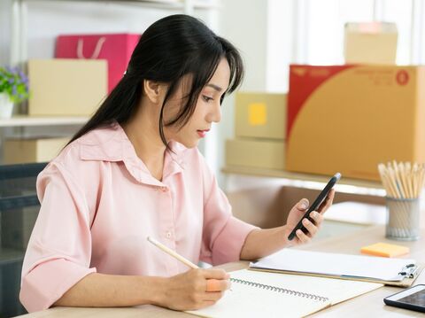 A woman is sitting on a chair and writing something in her notebook while watching her phone