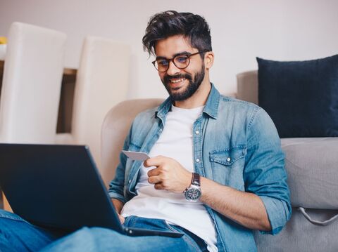 A young man with glasses and a beard, sitting comfortably in a living room and using a laptop. He holds a credit card, suggesting he is making an online purchase or managing financial transactions