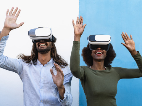 Two people enthusiastically experiencing virtual reality with VR headsets, raising their hands and smiling against a split white and blue background