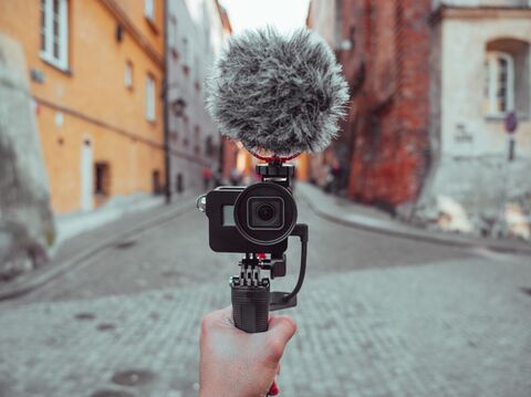 A hand holding a professional video camera equipped with a fluffy grey microphone on a stabilizer, recording a scenic cobblestone street in a charming old town