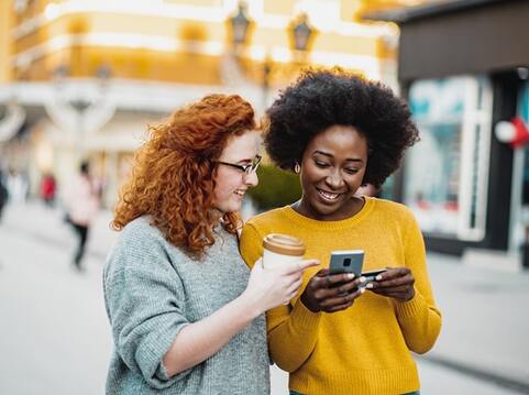 Two friends, one with curly red hair and the other with an afro, smiling and looking at a smartphone while enjoying coffee in an urban setting