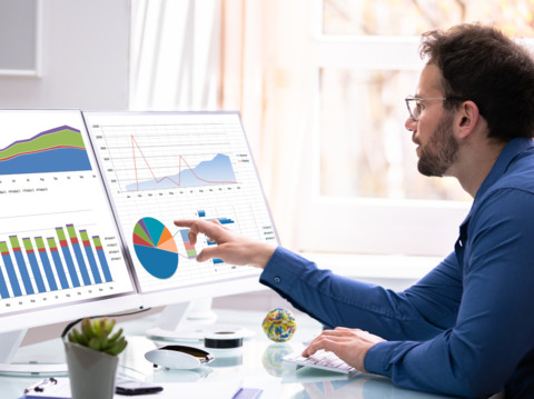 Man intensely analyzing data visualizations and graphs on two large computer monitors, focusing on performance metrics in a bright office setting