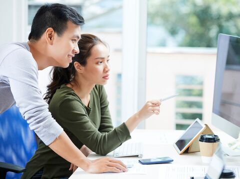 Two young professionals, a man and a woman, are focused on a computer screen in a brightly lit office. The man, wearing a light grey sweater, is standing and pointing at the screen with a pen, while the woman, seated in a green top, looks intently at the monitor