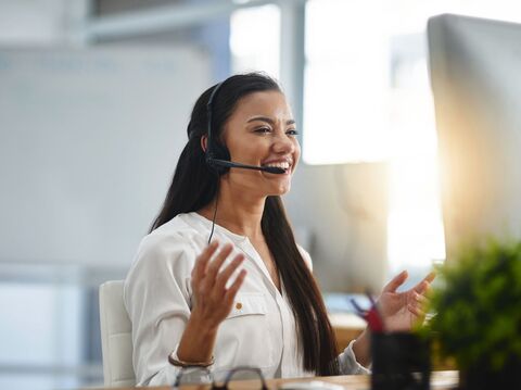 A woman wearing a headset with a microphone, seated on a chair and gesturing with her hand towards a monitor