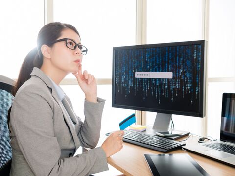 A woman in a gray blazer and glasses is seated at a desk, holding a blue credit card and looking thoughtfully at a computer screen. The screen displays a password entry field amidst a background of binary code. The setting indicates a focus on cyber security, possibly involving secure online banking or financial transactions