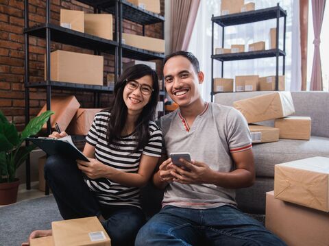 A man and woman are sitting on the floor in a modern home office filled with packaged boxes. The woman is holding a clipboard and writing, while the man holds a smartphone, likely managing logistics or customer service