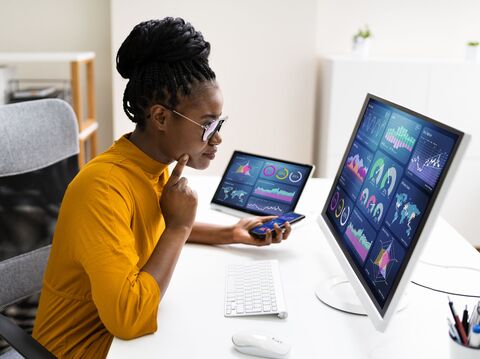 A woman sitting on a chair in front of her desktop, surrounded by graphs and data. She rests one finger on her cheek, indicating thoughtful data analysis