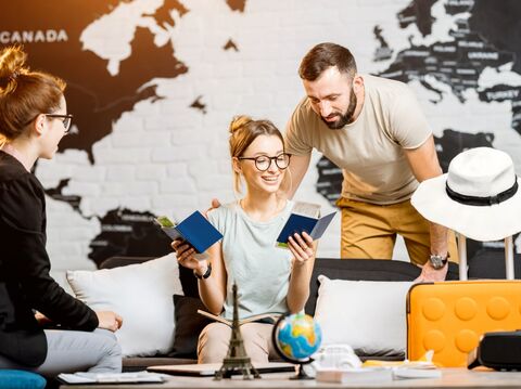 three people, two women and one man looking at their passports while having a packed suitcase with a fedora on the top of the handle and a background of a globe majorly focused on the north america continent