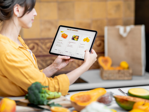 Woman using a tablet to browse an online grocery store while surrounded by fresh fruits and vegetables in her kitchen