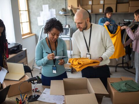 Two colleagues in a warehouse preparing clothing items for shipment, surrounded by boxes and packaging materials