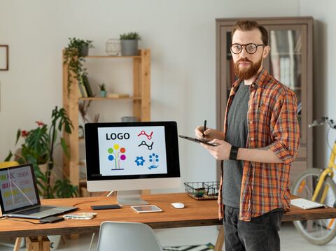 A graphic designer holding a tablet and pen, standing in a workspace with a computer displaying various logo designs