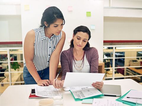 Two colleagues reviewing documents and discussing work in an office setting, with papers and notes spread out on the table