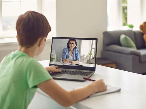 Child attending an online class, watching a lesson on a laptop while taking notes at home