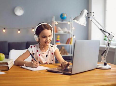 Young girl with headphones participating in an online class, smiling and taking notes while using a laptop