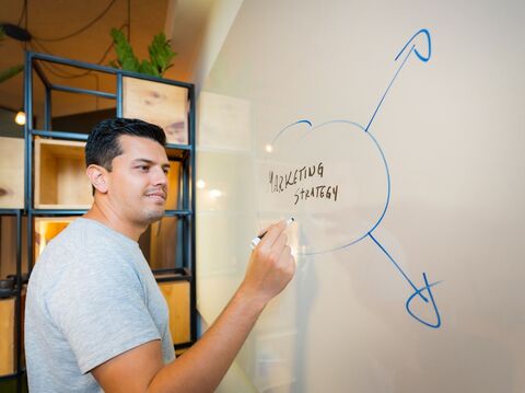 A man in a gray shirt is writing on a whiteboard. The board has \"Marketing Strategy\" written in the center with arrows pointing outward. The setting appears to be an office with a modern design, featuring shelves and plants in the background