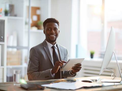 A professionally dressed Black man sitting at a desk reviewing documents on a digital tablet in a bright, modern office setting