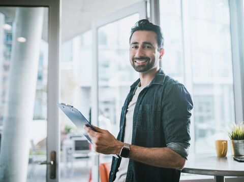 Smiling young man holding a clipboard in a modern office, showcasing a confident professional