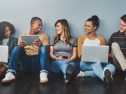 A group of three people are sitting on the floor. Some are holding laptops, some are holding phones, and some are looking at each other