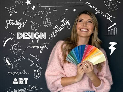 A woman is standing in front of a black wall and holding some colorful cards. The wall is full of words like \"design,\" \"creativity,\" \"ar, \"Creativity\" , \"Art\", \"Concept, etc\"