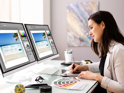 A professional woman graphic designer working on two computer monitors with graphic design software open, showing beach-themed layouts; her workspace is neat with design samples and tools