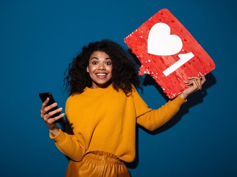 A woman in a yellow dress, holding a smartphone in one hand and a social media like in another hand