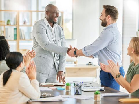 In a meeting room, two men are shaking hands, and the other is clapping