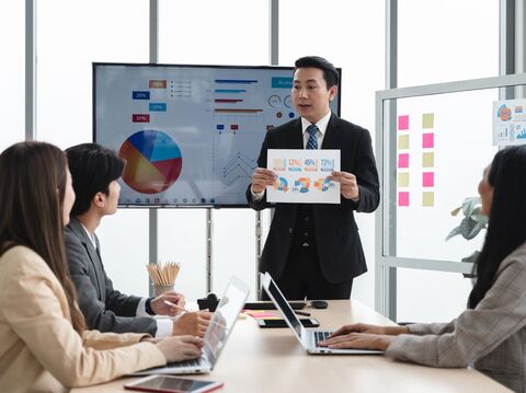 In a modern corporate meeting room, a well-dressed businessman is presenting statistical data to a group of three attentive colleagues. He holds a colorful infographic handout and stands beside a large digital display showing pie charts and graphs, indicating business performance metrics. His audience, two women and one man, are dressed in business attire and are engaged, taking notes and looking towards the speaker