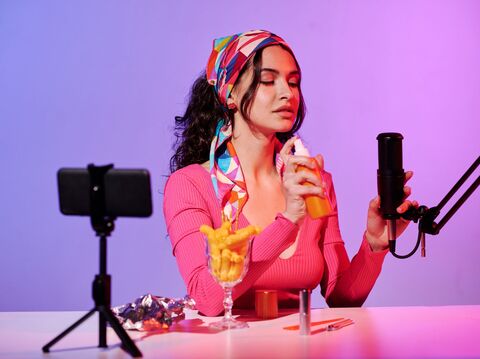 A woman with long curly hair, wearing a colorful headscarf and a pink top, is recording content. She is holding a spray bottle in one hand and a microphone in the other. There is a smartphone on a tripod, a glass filled with snacks, and some tools on the table in front of her