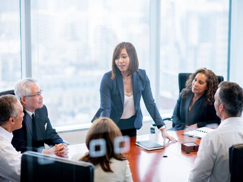 A young woman leading a meeting with diverse colleagues in a modern office overlooking a city. The group, attentively engaged, sits around a conference table with digital devices