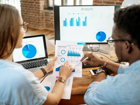 Two colleagues analyzing data charts and graphs displayed on a laptop and a monitor