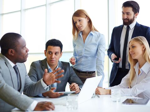 The image shows a diverse group of five business professionals engaged in a meeting around a table in a bright office space. A man gestures as he speaks, holding the attention of his colleagues. Two women and three men, all dressed in business attire, listen intently and discuss ideas