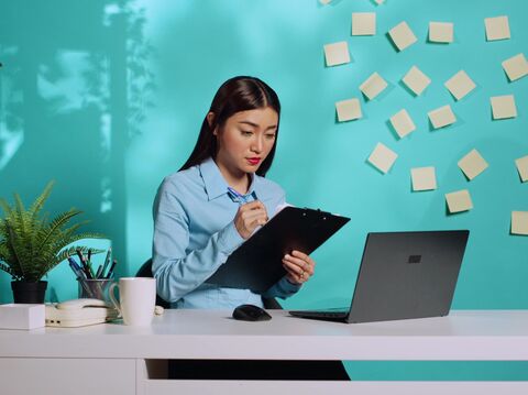 A young woman reviewing notes and working on a laptop in a modern office with a green backdrop