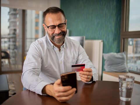 A middle-aged man smiling while using a smartphone and holding credit cards, sitting in a contemporary office