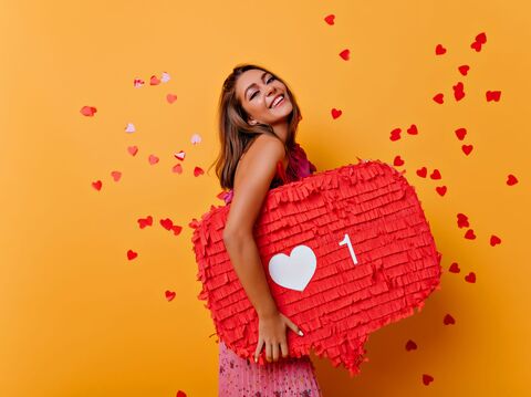 A cheerful woman in a pink dress stands against a vibrant orange background, holding a large red heart-shaped piñata with a white heart icon and the number '1'. Red heart confetti surrounds her, creating a lively and celebratory atmosphere