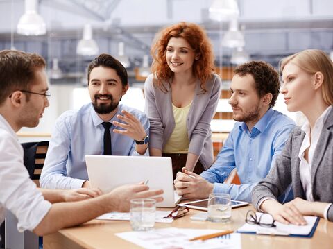 A group of diverse young professionals engaged in a collaborative meeting around a laptop in a modern office