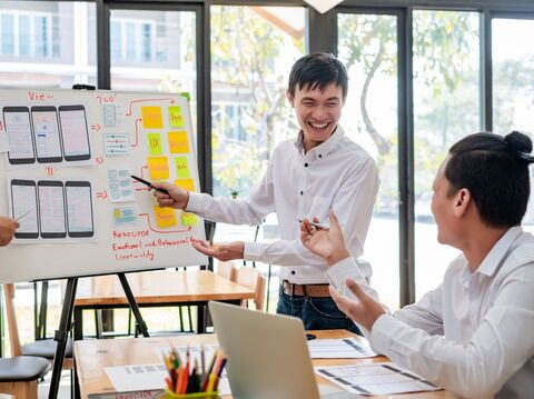 In a meeting room, a person is explaining something on a whiteboard while others are listening to him