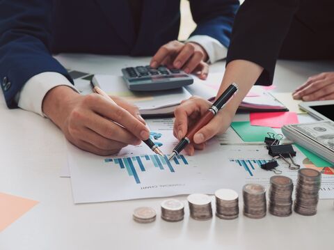 Close-up of two professionals' hands pointing at financial charts on a table, with stacks of coins and a calculator