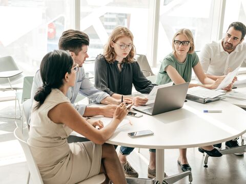 A diverse group of five people gathered around a round table, engaged in a discussion. One woman is holding a document, while a man points at a laptop screen
