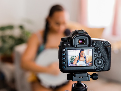 A focused woman being filmed by a digital camera on a tripod, reviewing content on the camera’s display screen